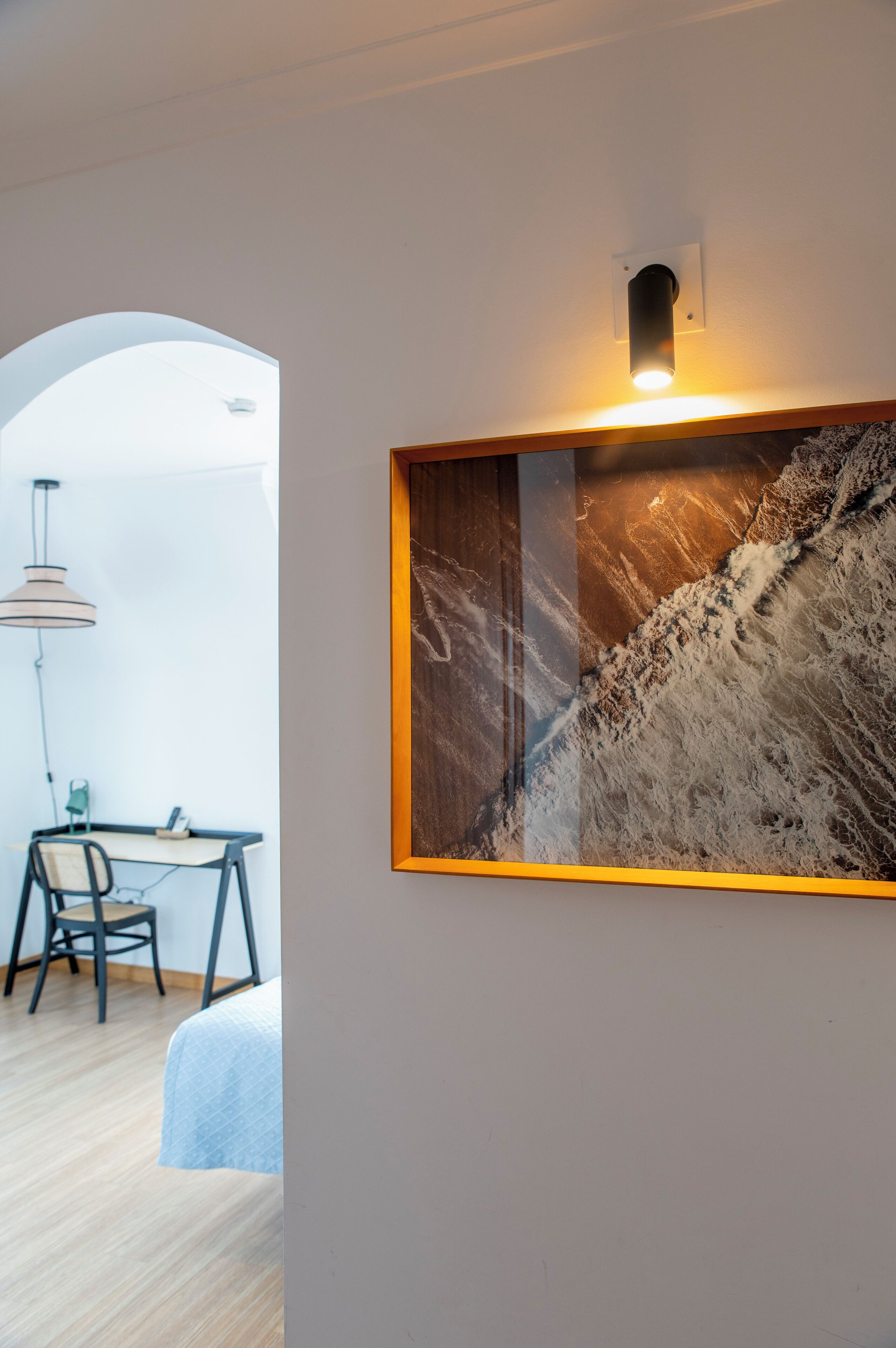 Una habitación con una cama, un escritorio con una silla, una lámpara colgante y un cuadro abstracto enmarcado.