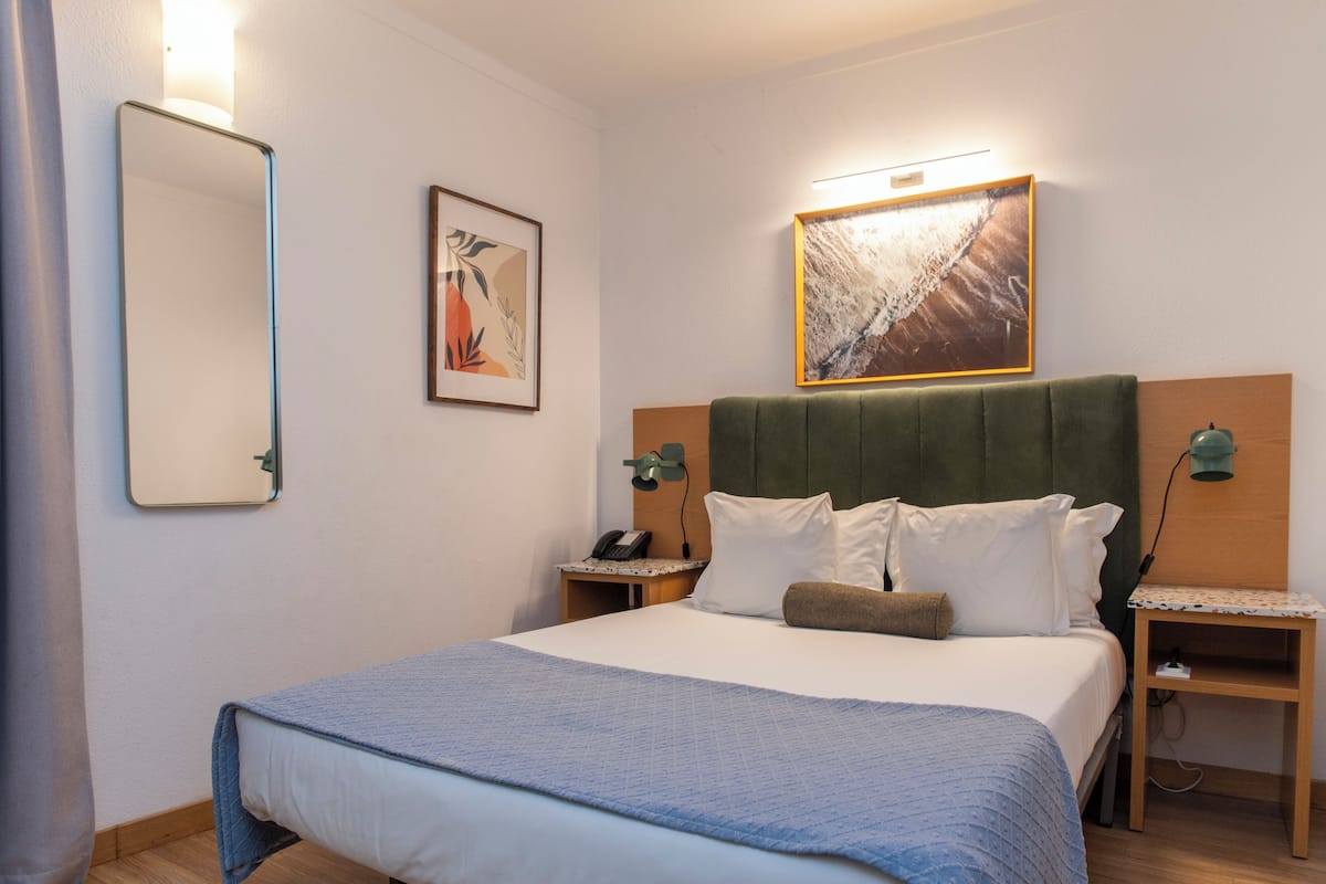Een keurig opgemaakte hotelkamer met een groot bed, twee nachtkastjes, een wandspiegel, een ingelijst kunstwerk en een wandlamp.