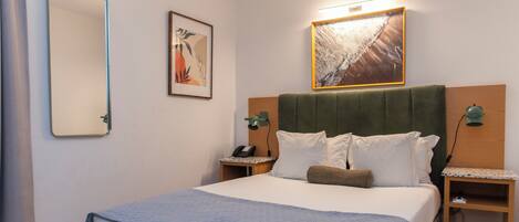 A neatly arranged hotel room with a large bed, two bedside tables, a wall-mounted mirror, a framed artwork, and a wall-mounted lamp.