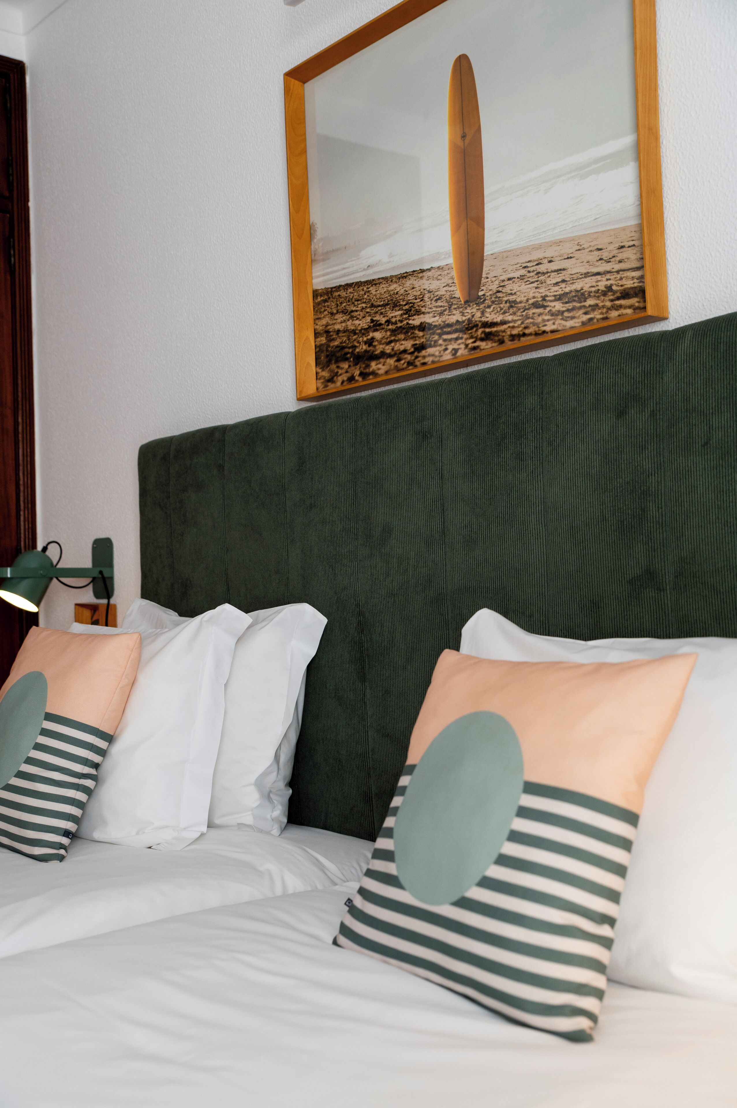 Una cama bien hecha con ropa blanca y almohadas a rayas, un cabecero tapizado en verde y un cuadro con una tabla de surf en la pared.