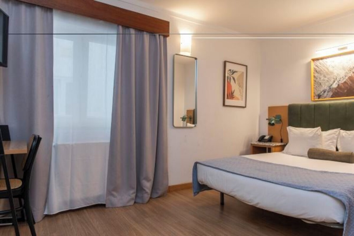 A hotel room with a bed, a desk, a chair, a nightstand, and a mirror.