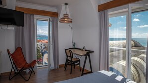 Una habitación con cama, escritorio, silla y televisor. Hay un ventanal con vistas al mar y a edificios.