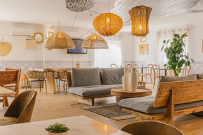 Un salon chaleureux et bien éclairé, avec un canapé, des fauteuils et une table à manger. On y trouve des lampes suspendues en osier et une grande plante dans un coin.