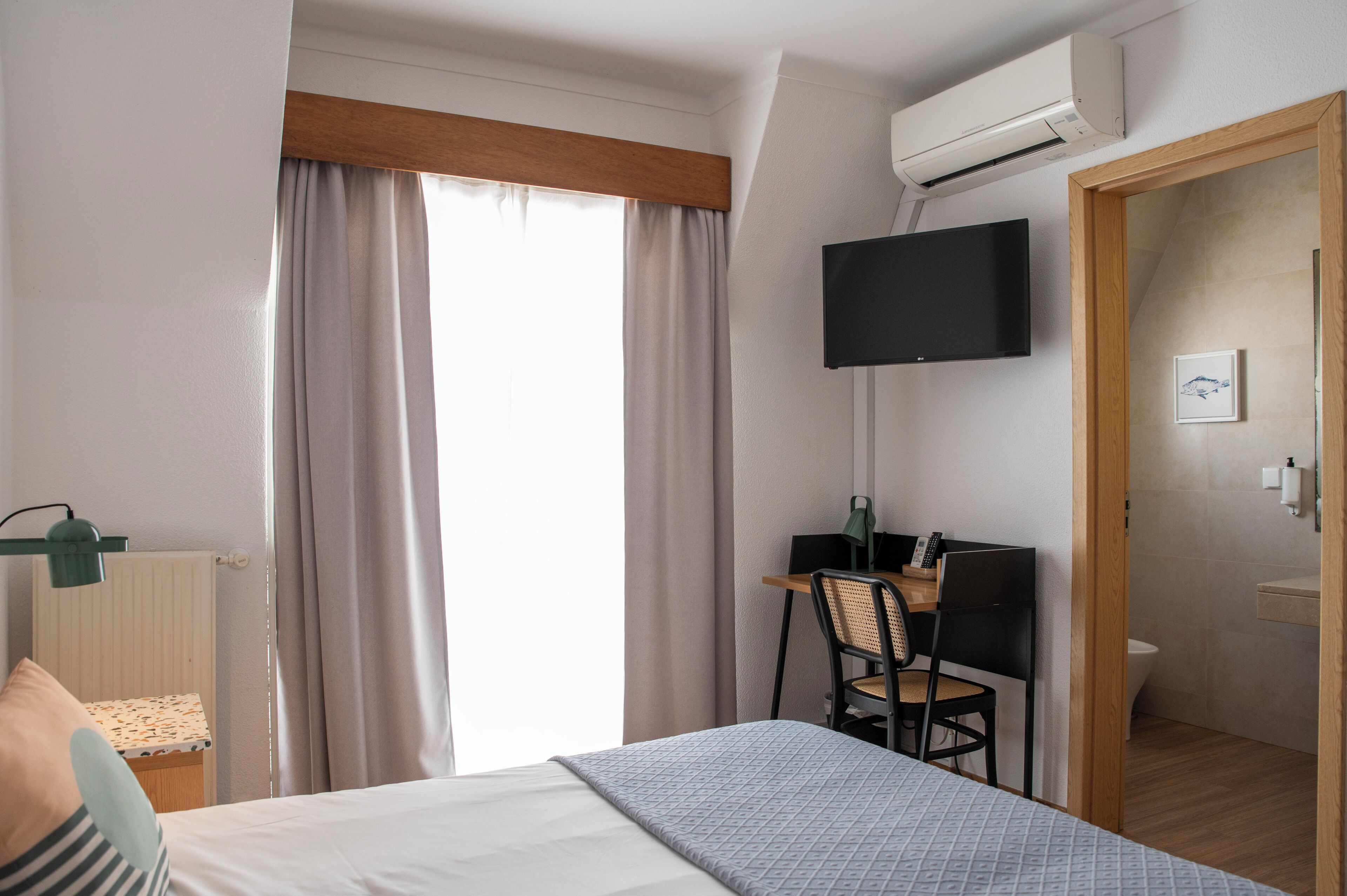 A hotel room with a bed, a desk with a chair, a television mounted on the wall, and a bathroom visible through an open door.