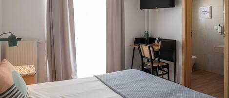A hotel room with a bed, a desk with a chair, a television mounted on the wall, and a bathroom visible through an open door.