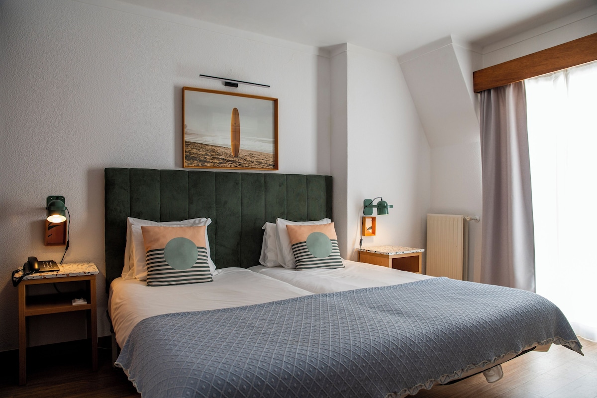 Una cama doble con cabecera verde, dos mesitas de noche con lámparas, un cuadro enmarcado sobre la cama y una ventana con cortinas.