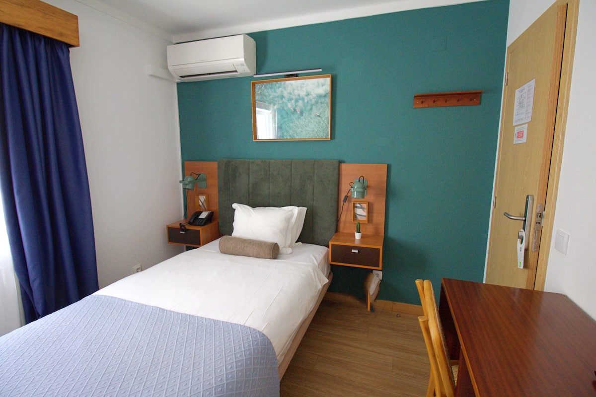 Een hotelkamer met een bed, nachtkastjes, een airconditioner, een schilderij en een houten deur.