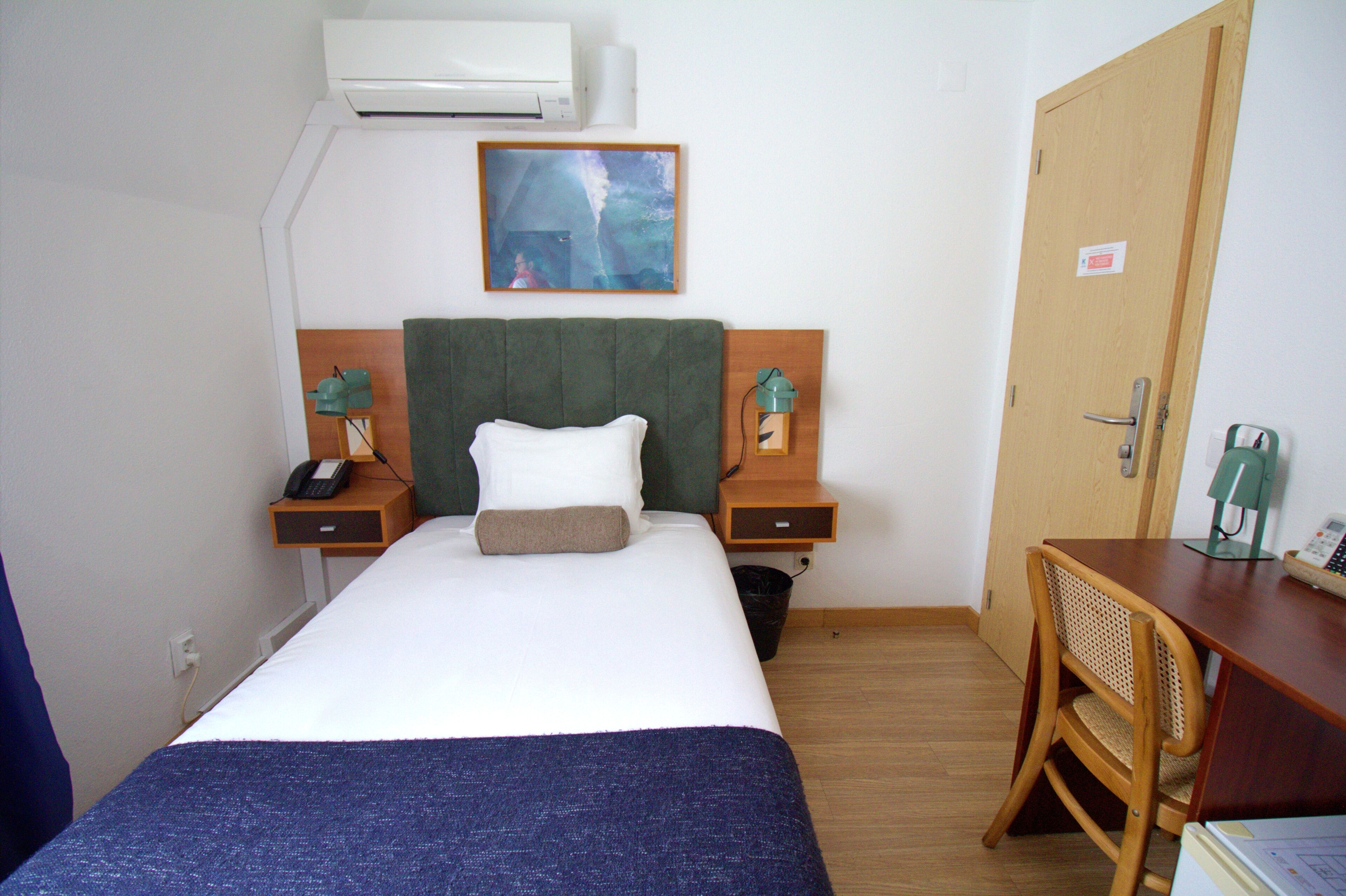 A hotel room with a bed, a desk, a chair, a nightstand with a phone, a lamp, and an air conditioning unit.