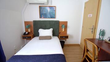 A hotel room with a bed, a desk, a chair, a nightstand with a phone, a lamp, and an air conditioning unit.