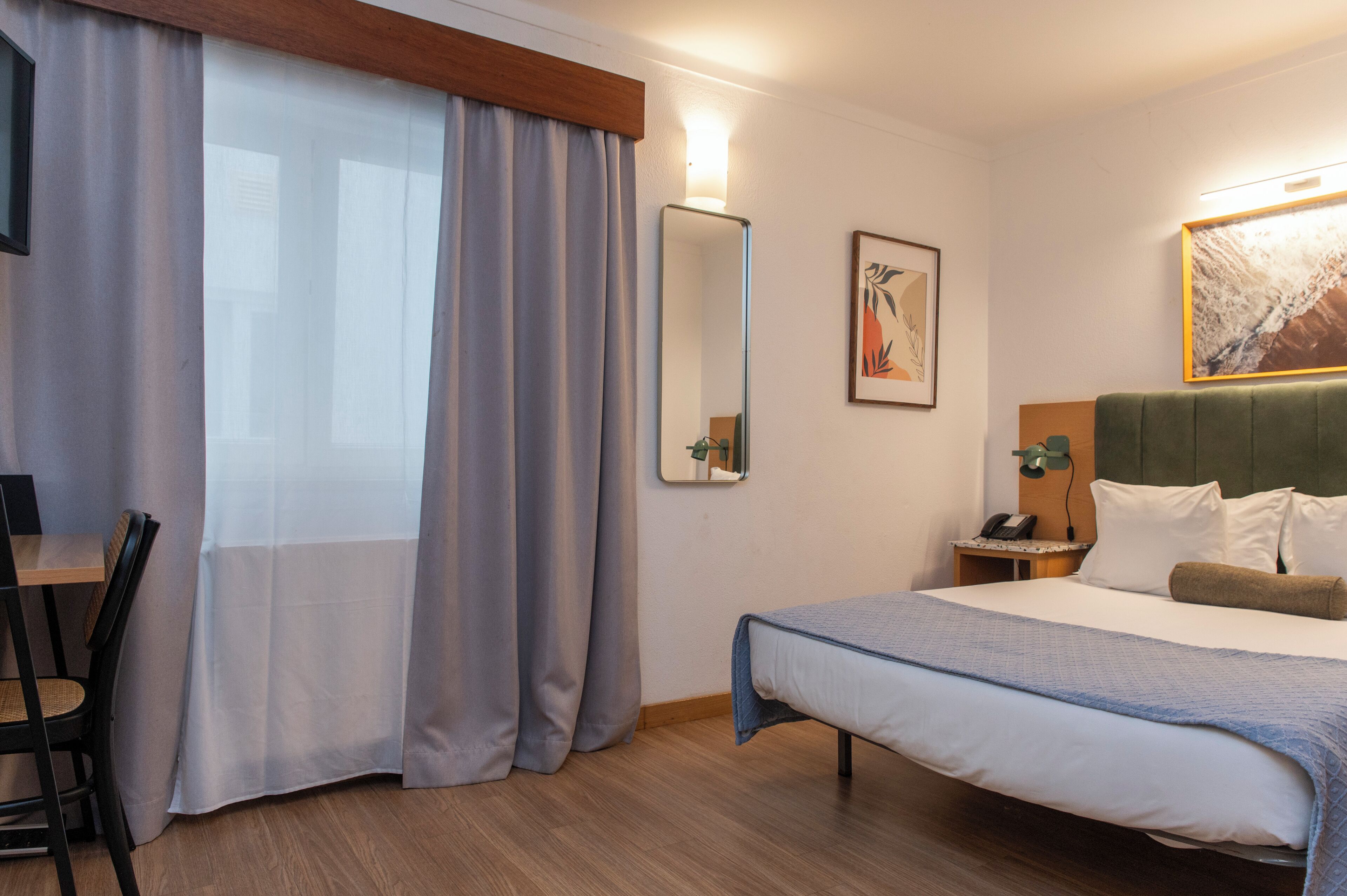 Habitación de hotel con cama, escritorio, silla, mesita de noche, espejo y obras de arte en las paredes.