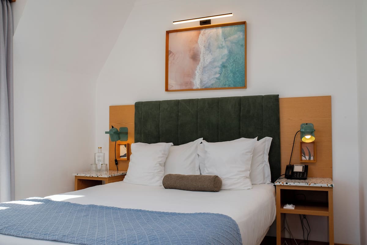 Een net opgemaakt bed met een blauwe deken, een houten hoofdbord en een schilderij van een strandlandschap erboven.