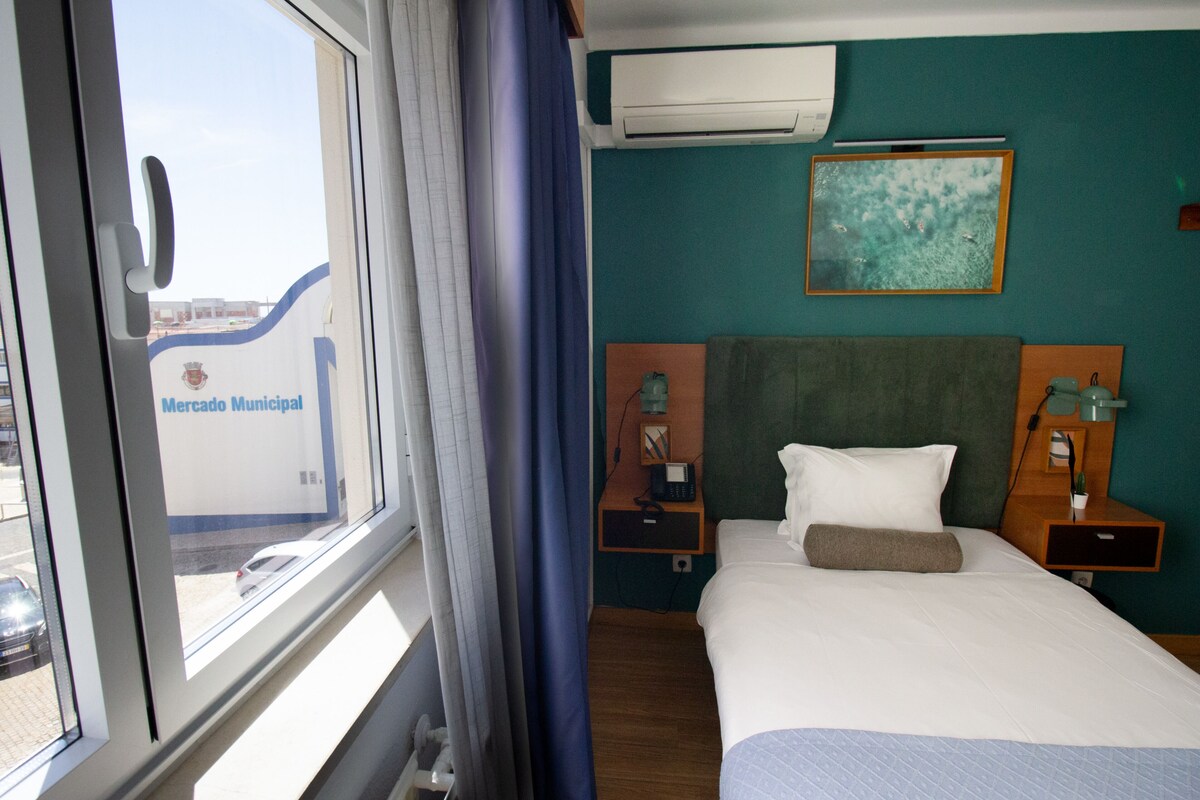 Habitación de hotel con cama, mesita de noche, lámpara, equipo de aire acondicionado y ventana con cortinas.
