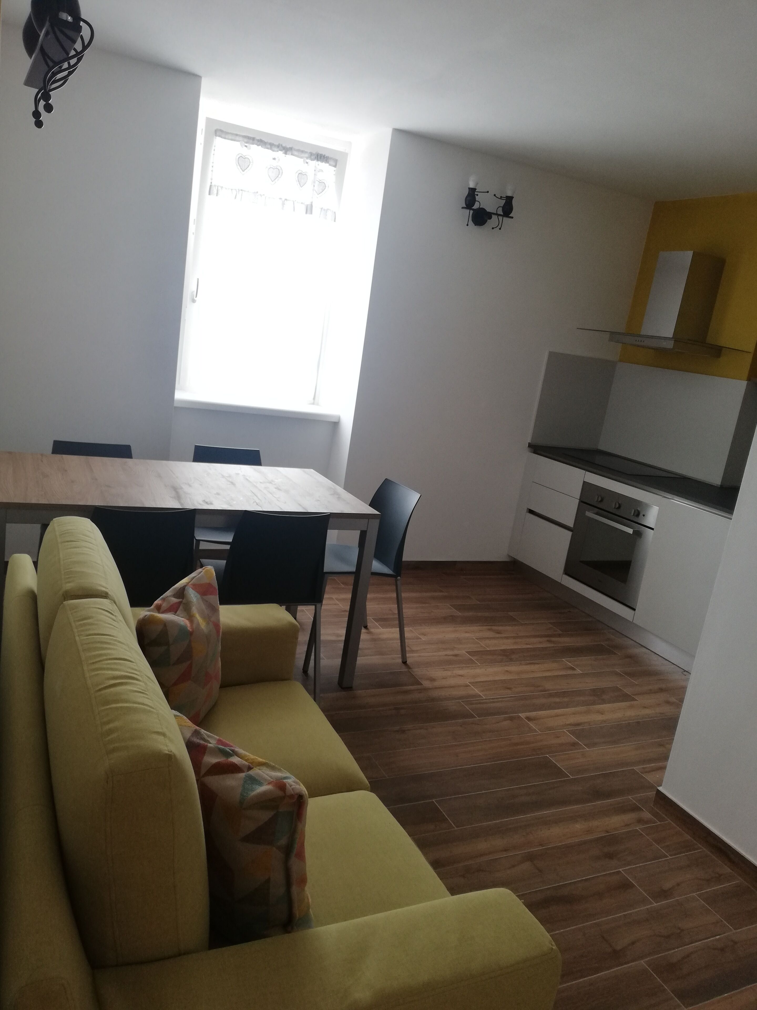 A modern living room with a sofa, a dining table, and chairs. There is a kitchen area with a stove and cabinets.