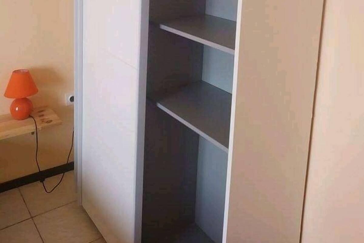 Quarto com um guarda-roupa branco, uma pequena luminária laranja sobre uma mesa de cabeceira de madeira e um chão ladrilhado.