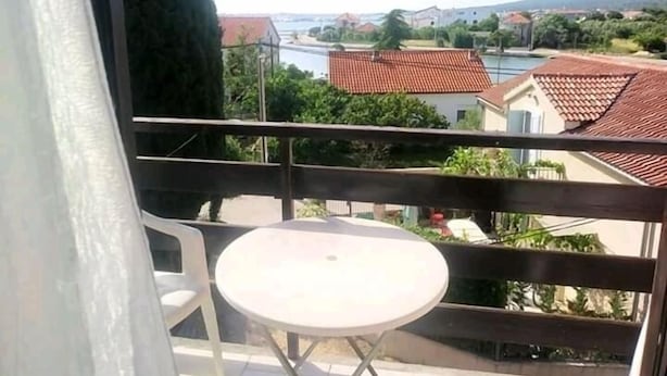 A balcony with a white table and chairs, overlooking a residential area with red-tiled roofs and greenery.