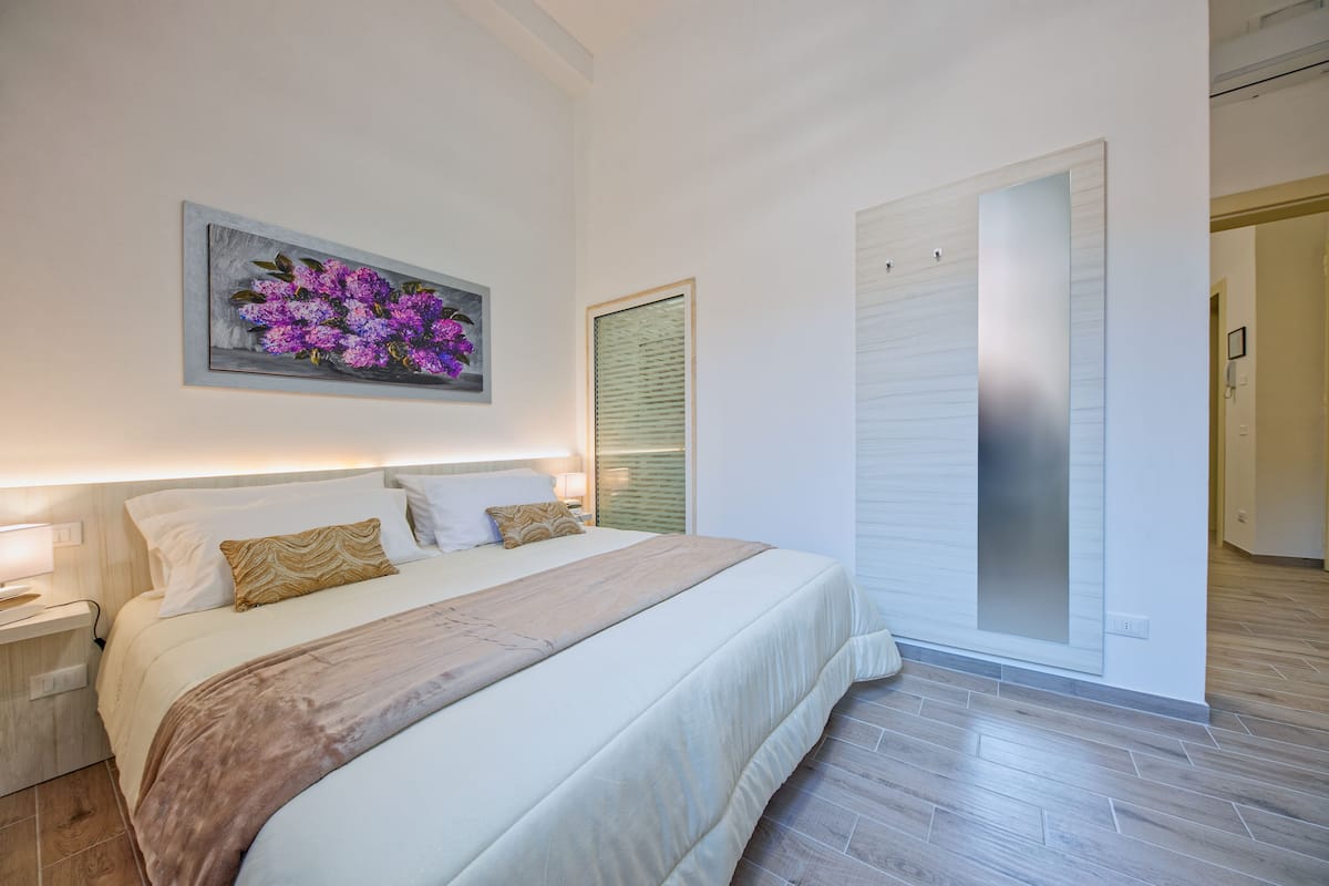 A neatly arranged bedroom with a large bed, a bedside table, a wall-mounted artwork of purple flowers, and a sliding door.