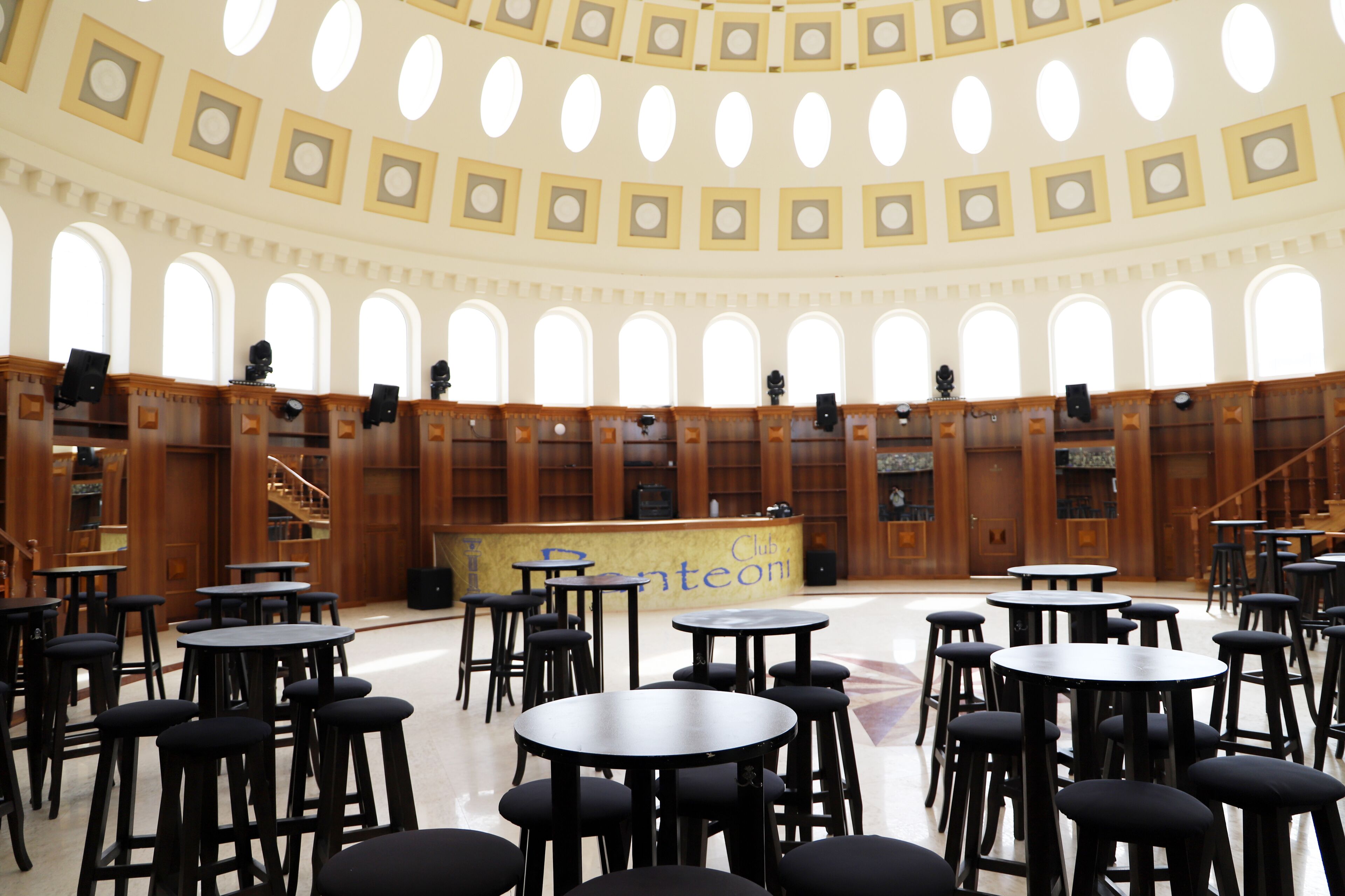 A library with multiple tables and chairs, a central counter, and a staircase leading to upper levels.