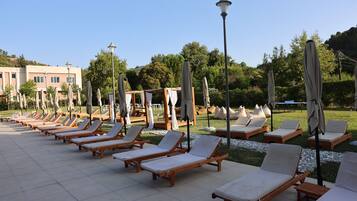 A row of wooden lounge chairs with white cushions, arranged on a tiled patio with a lamp post and umbrellas in the background.