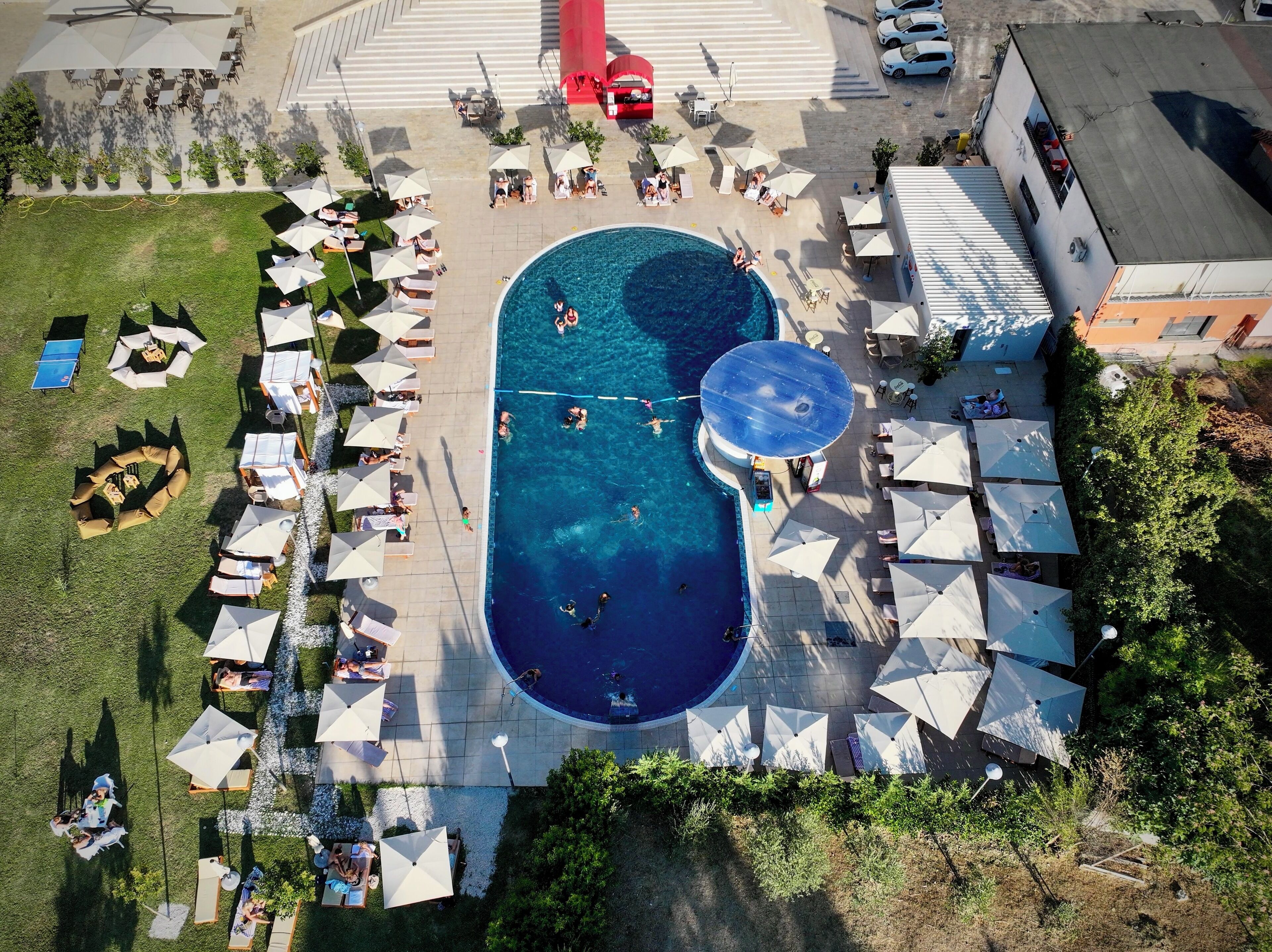 An aerial view of a pool area with multiple lounge chairs and umbrellas, surrounded by greenery and a building.