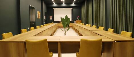Een vergaderruimte met een lange tafel en gele stoelen, een projectiescherm en een grote plant in het midden.