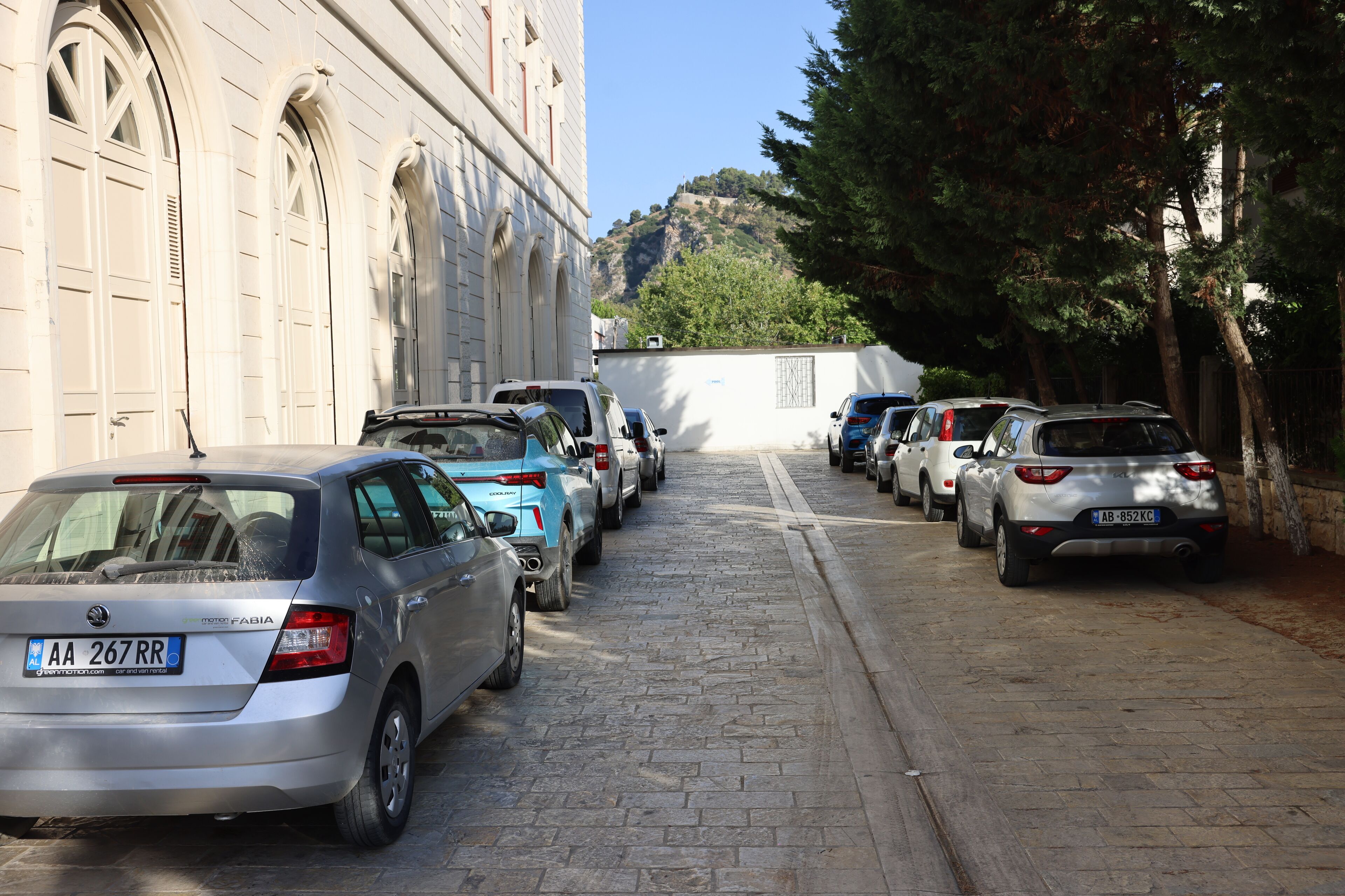 A row of parked cars along a cobblestone street with a building featuring arched windows on the left.