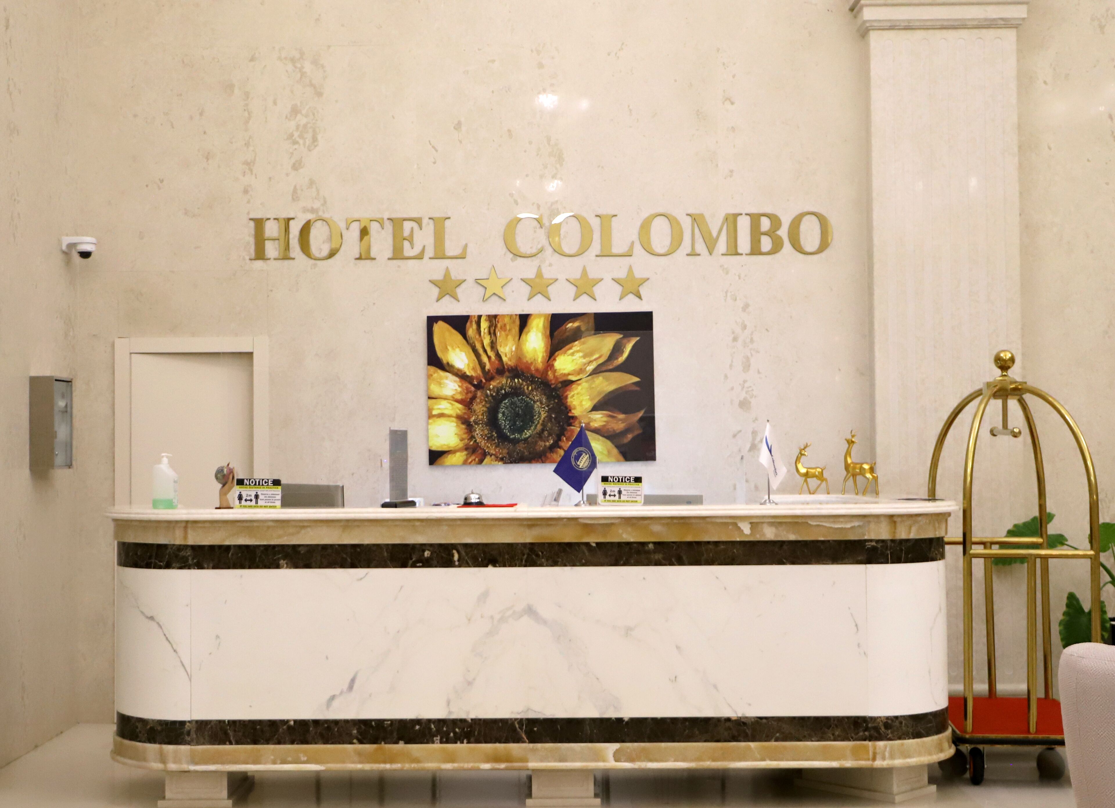 Mostrador de recepción del Hotel Colombo con encimera de mármol, una obra de arte con girasoles y un cartel que indica una calificación de 5 estrellas.