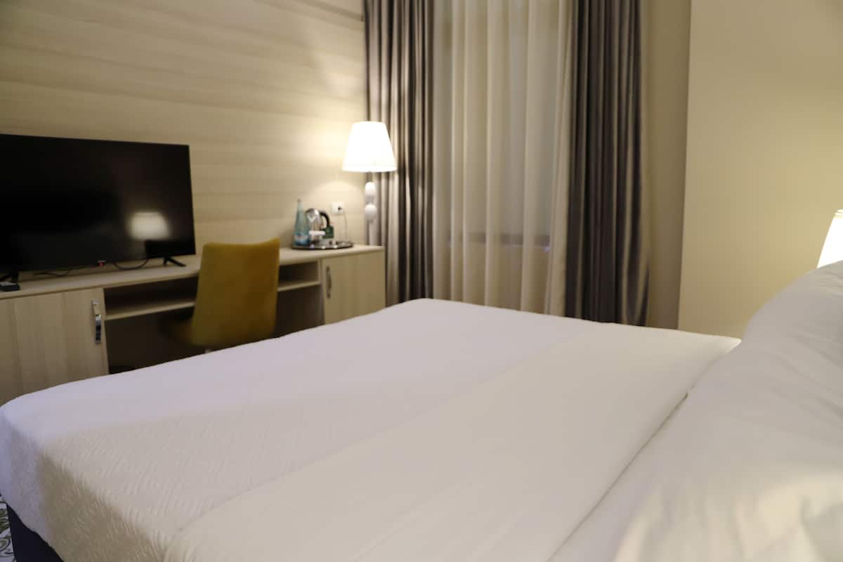 Quarto de hotel com uma cama grande, uma escrivaninha com cadeira, televisão e luminária.
