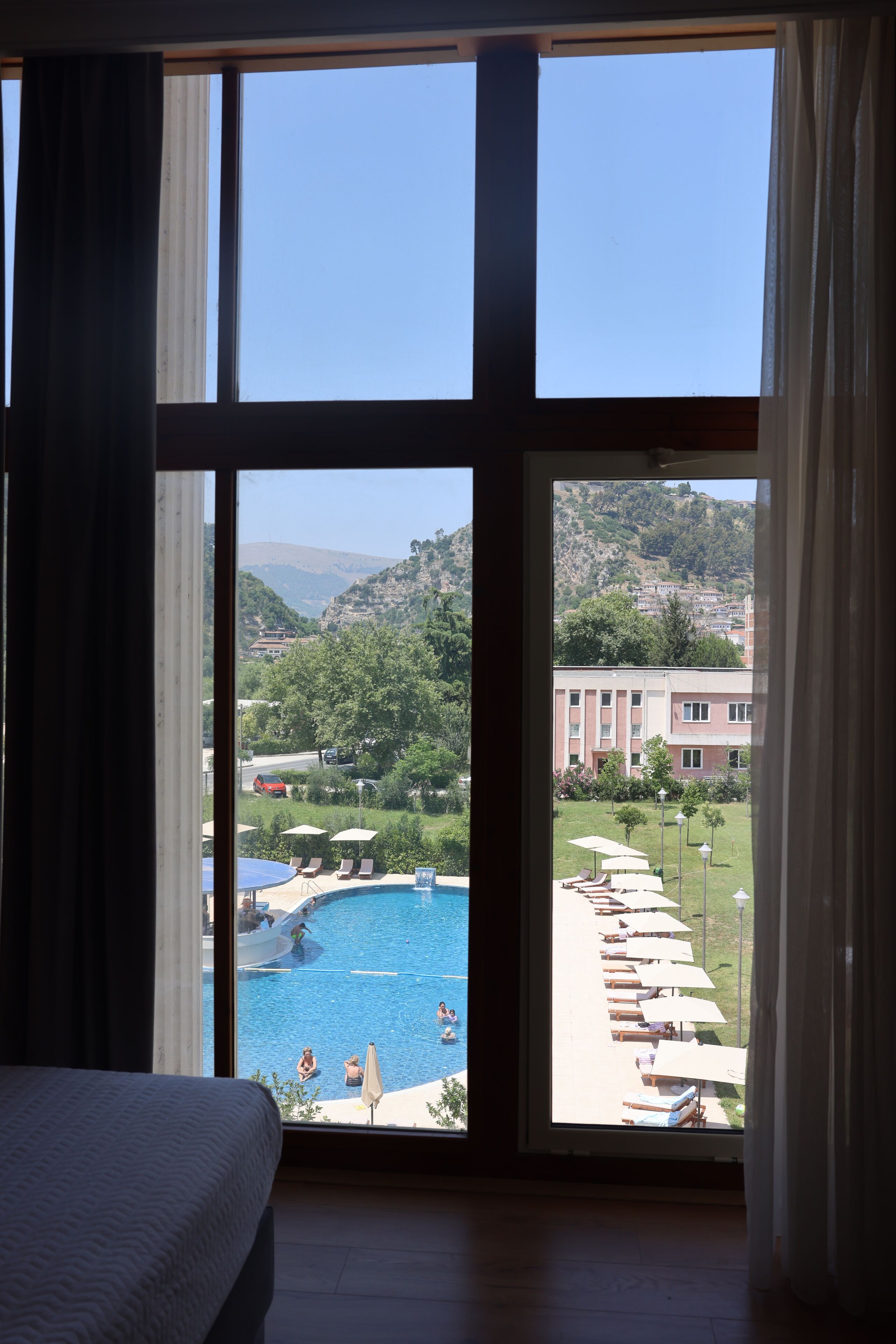 A hotel room with a large window overlooking a swimming pool and a mountainous landscape.