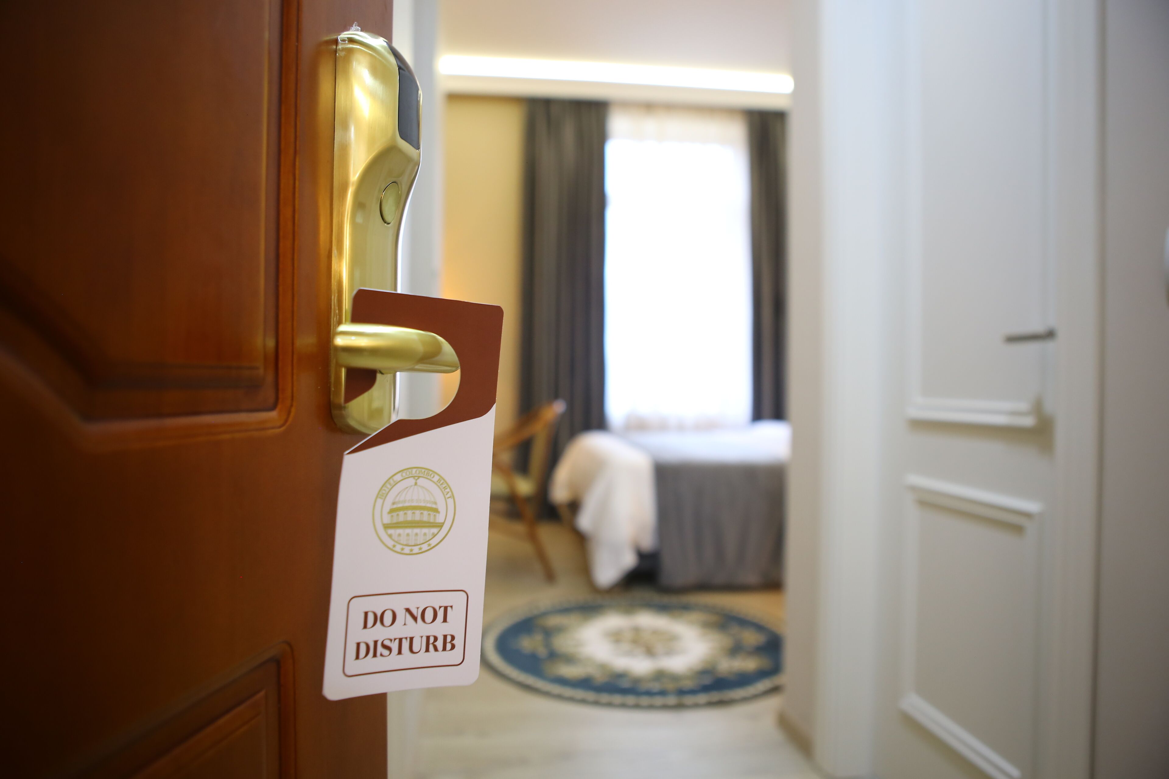 A hotel room with a bed, a chair, and a rug. A sign on the door reads "DO NOT DISTURB".