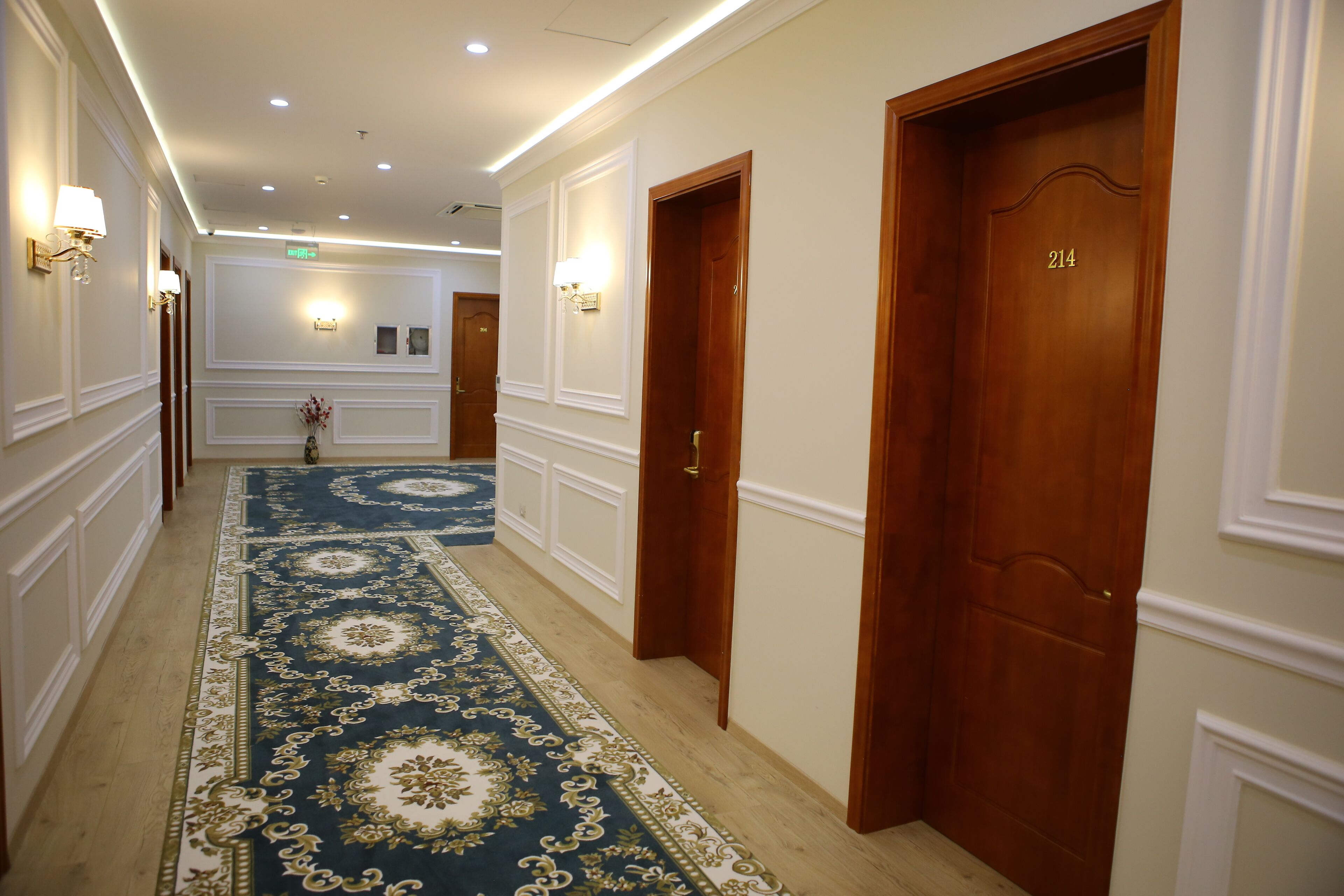 A hotel hallway with a blue and gold patterned carpet, wooden doors, and wall-mounted light fixtures.