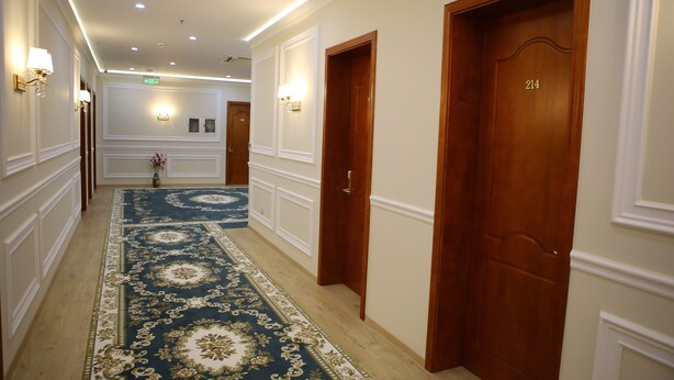 Un pasillo de hotel con alfombra de diseño azul y dorado, puertas de madera y luminarias fijadas a la pared.