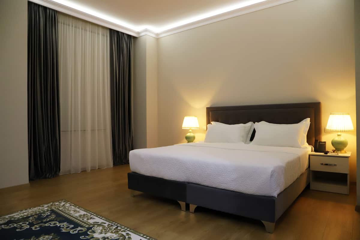 Cama bem arrumada com lençóis brancos, duas luminárias de cabeceira, mesa de cabeceira e uma janela grande com cortinas.
