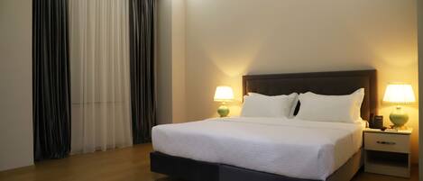 Una cama bien hecha con sábanas blancas, dos lámparas de noche, una mesita de noche y un ventanal con cortinas.