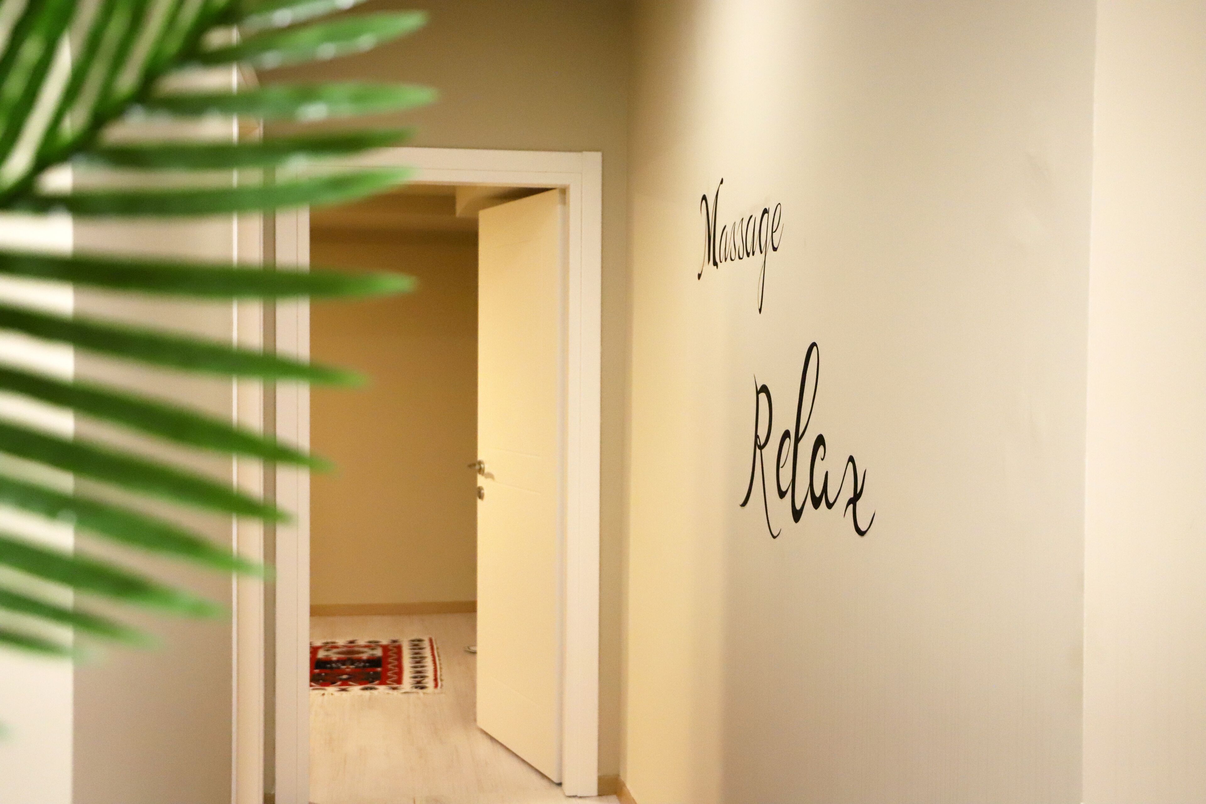 A hallway with a door leading to a room, a wall with the word "Massage" and "Relax" written on it, and a plant in the foreground.