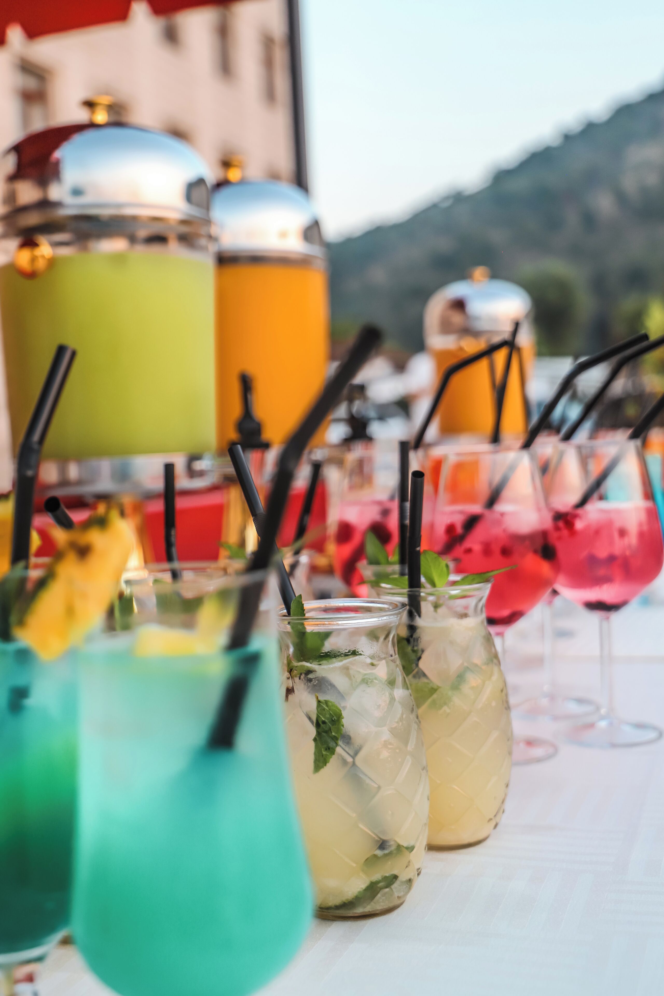 A row of colorful drinks with straws and mint leaves, set against a backdrop of a building and mountains.