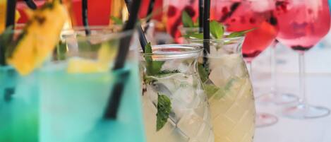A row of colorful drinks with straws and mint leaves, set against a backdrop of a building and mountains.