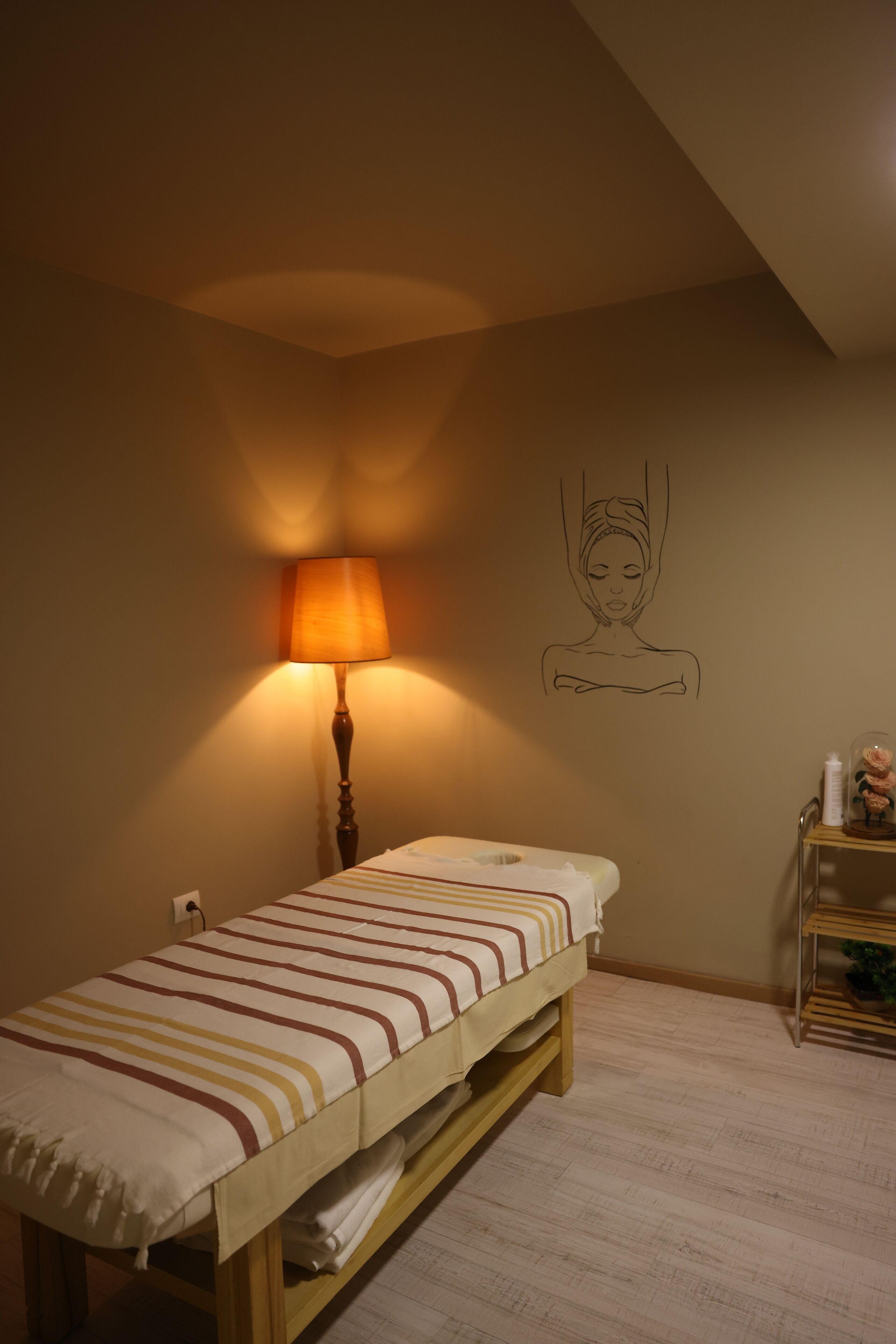 Una cama individual con ropa de cama a rayas, una lámpara de noche, una obra de arte colgada en la pared y una mesita con una botella y un objeto decorativo.