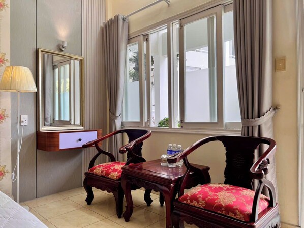 Habitación de hotel con cama, escritorio con lámpara, dos sillas con cojines rojos, una mesita con botellas de agua y una ventana con cortinas.