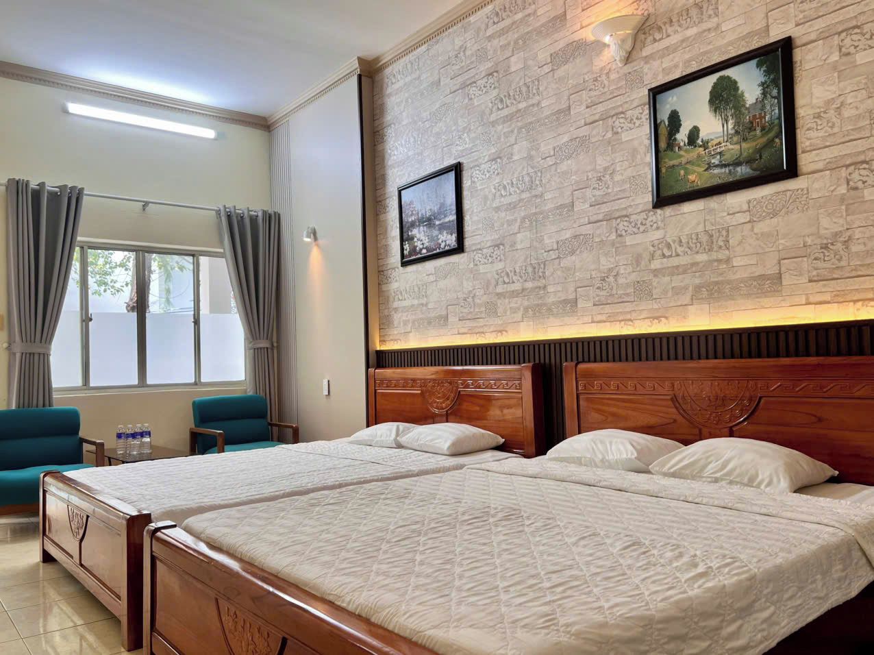 A hotel room with two double beds, a wooden headboard, a small table with water bottles, a blue armchair, and a window with curtains.