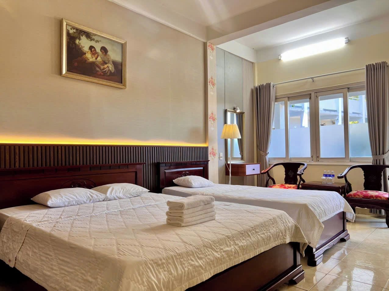 A hotel room with two beds, a painting on the wall, a lamp, and a view of the outside through the window.