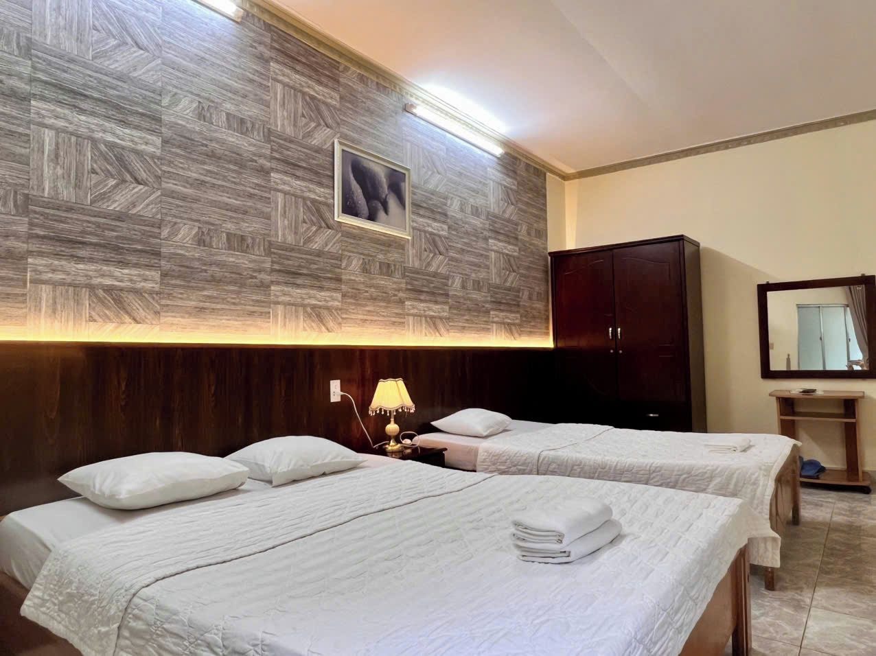 A hotel room with two beds, a wooden headboard, a nightstand with a lamp, a wardrobe, a mirror, and a wall-mounted picture.