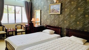 A hotel room with two beds, a desk with a chair, a window with curtains, and a wall with a patterned wallpaper.