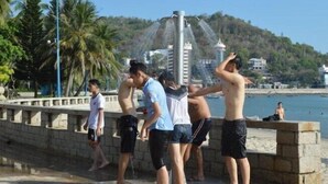A group of people standing by a water feature on a waterfront promenade with a view of the sea and buildings in the background.