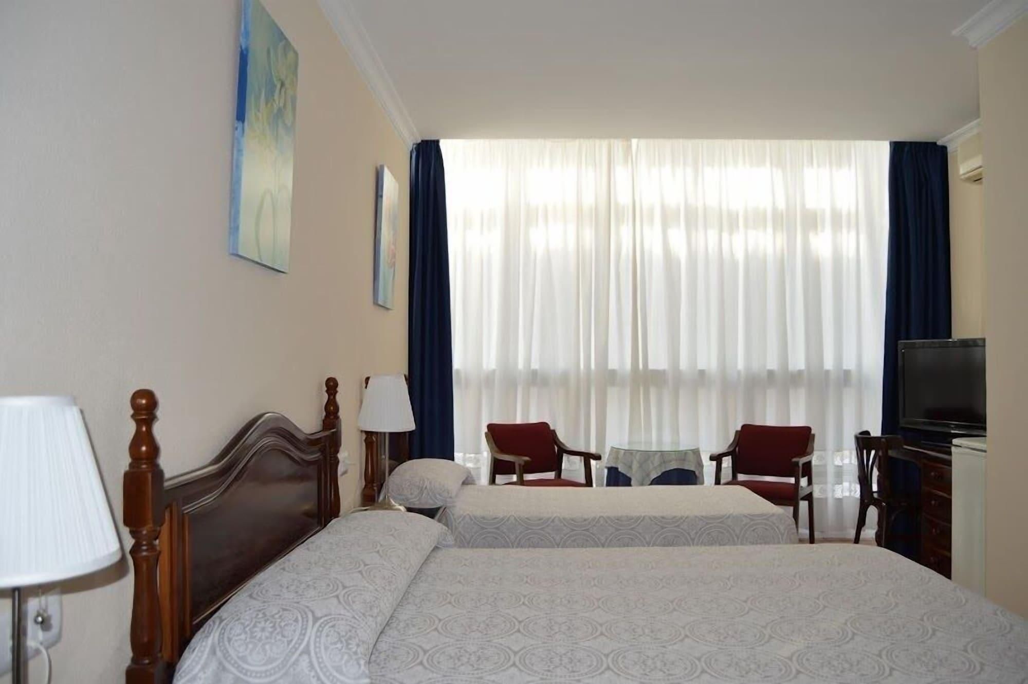 Habitación de hotel con dos camas, un escritorio, sillas, un televisor y una ventana con cortinas.