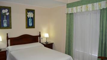 Una habitación de hotel con una cama individual, dos mesitas de noche con lámparas, una cortina verde y dos cuadros florales en la pared.