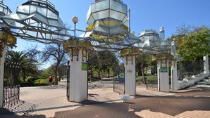A gated entrance with a geometric glass structure, paved walkway, and surrounding greenery.