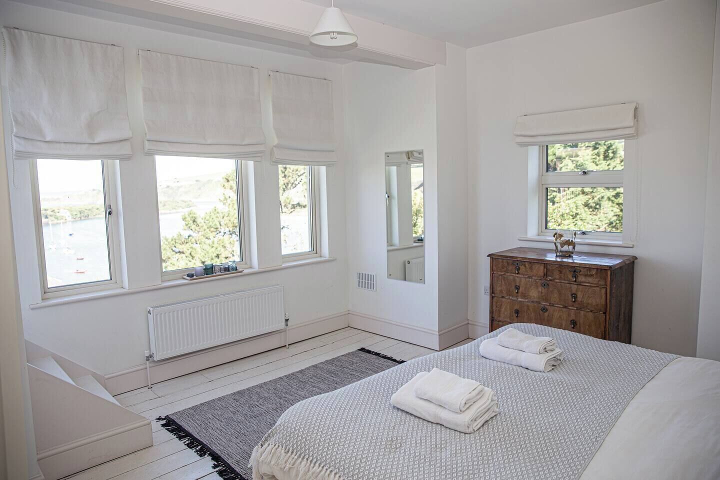 A bedroom with a bed, a dresser, a mirror, and three windows with white curtains.
