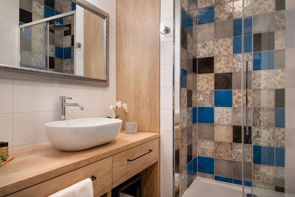 A bathroom with a large white vessel sink, a wooden vanity, a mirror, and a shower with blue and beige tiles.