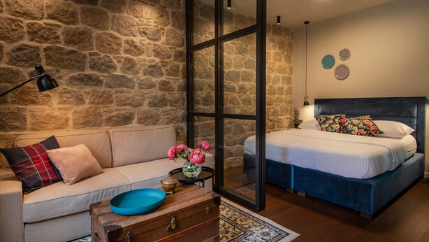 Un dormitorio con pared de piedra, una cama con cabecera oscura, un sofá con cojín escocés, un tazón azul sobre un cofre de madera, y un florero con flores rosadas.