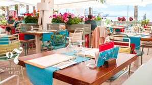 Un restaurante al aire libre con mesas y sillas, algunas con sombrillas, y vista al mar y barcos en el fondo.